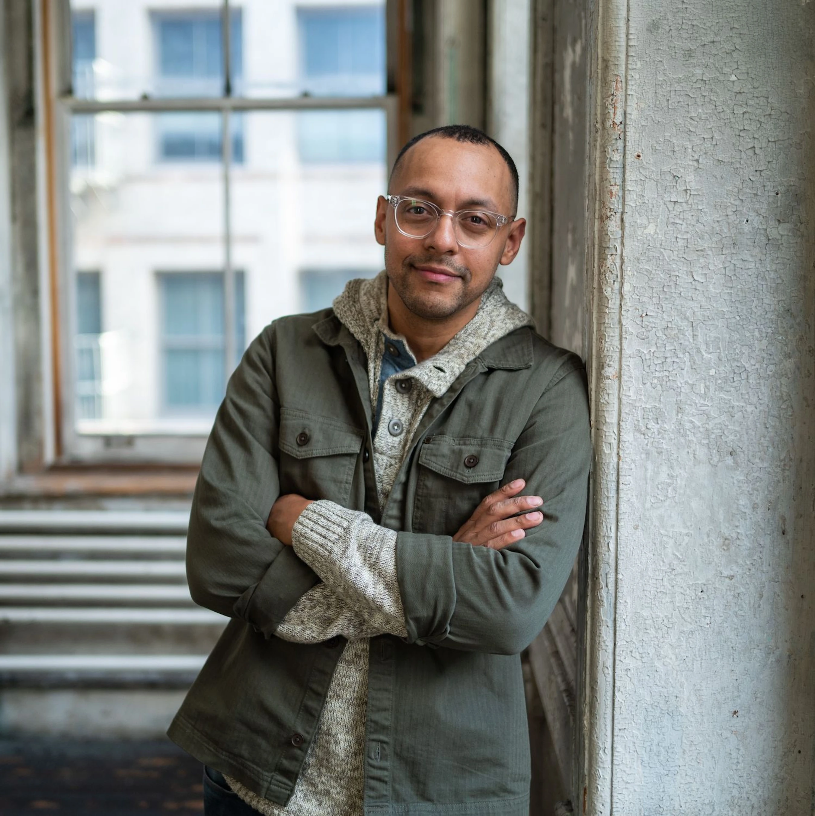 A man wearing glasses, a green jacket, and a sweater leans against a wall with peeling paint. He has his arms crossed and is looking at the camera.